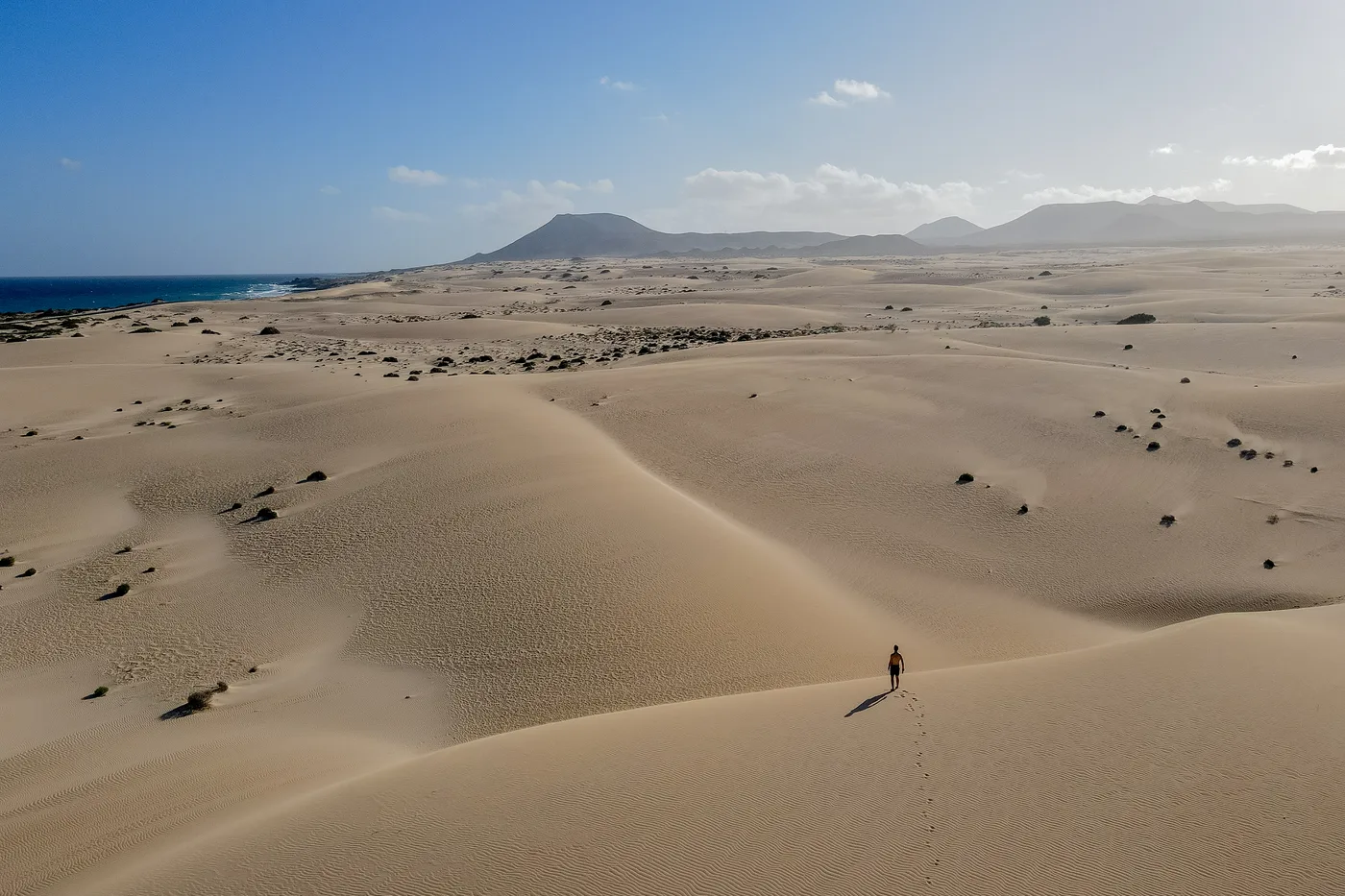 A lone walker crossing a high dune ridge in the Parque Natural de Corralejo, with the Atlantic and Fuerteventura's inland peaks in the distance