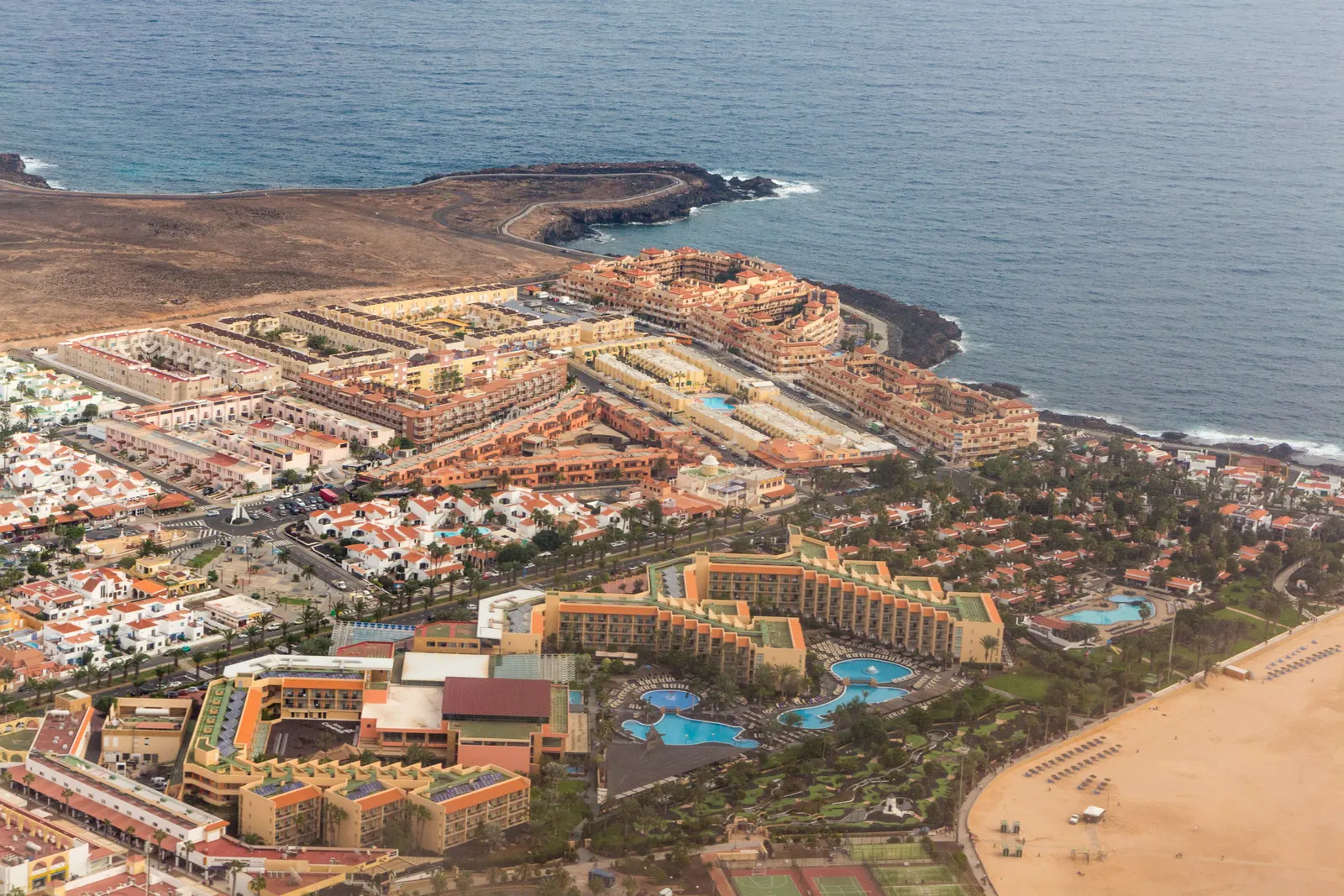 Aerial view of the horseshoe bay at Caleta de Fuste, with hotels and apartment blocks along the shore, a small rocky headland and the beach running along the water's edge