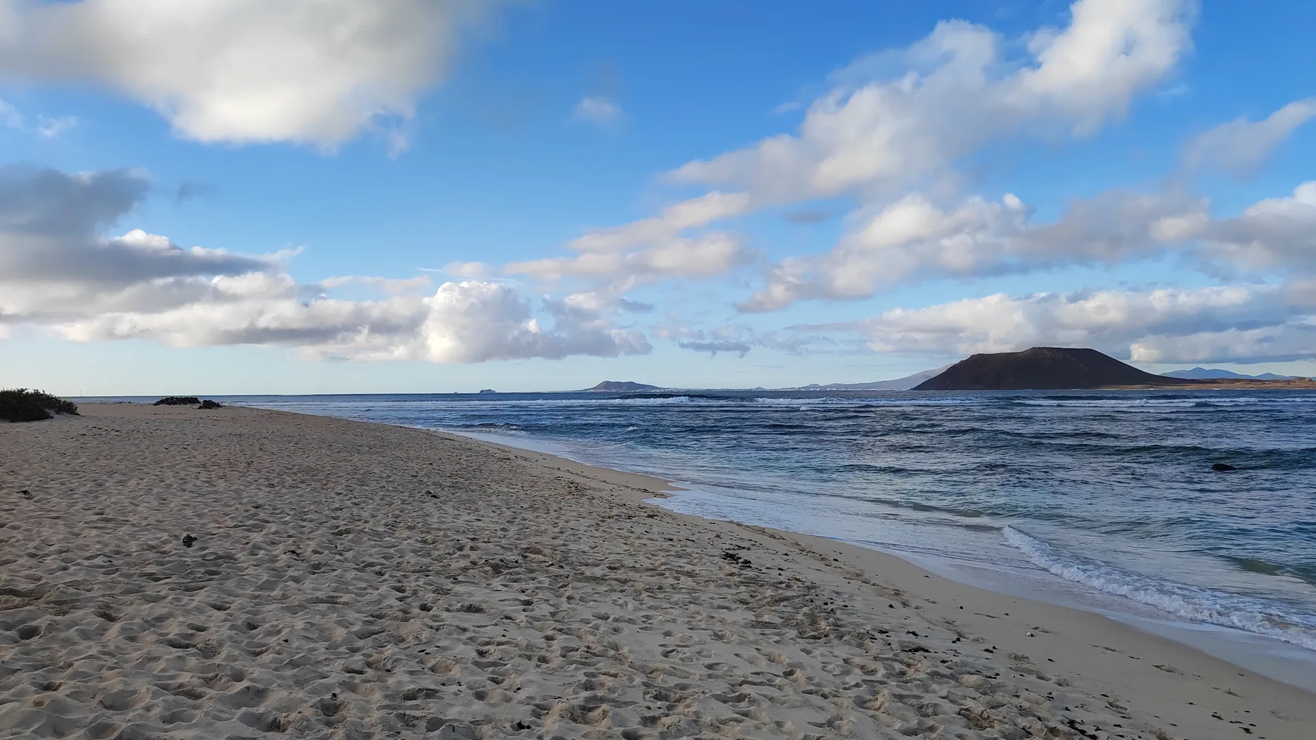 A quiet stretch of golden sand at Playa del Médano with gentle waves and a dark volcanic hill visible across the water in the distance