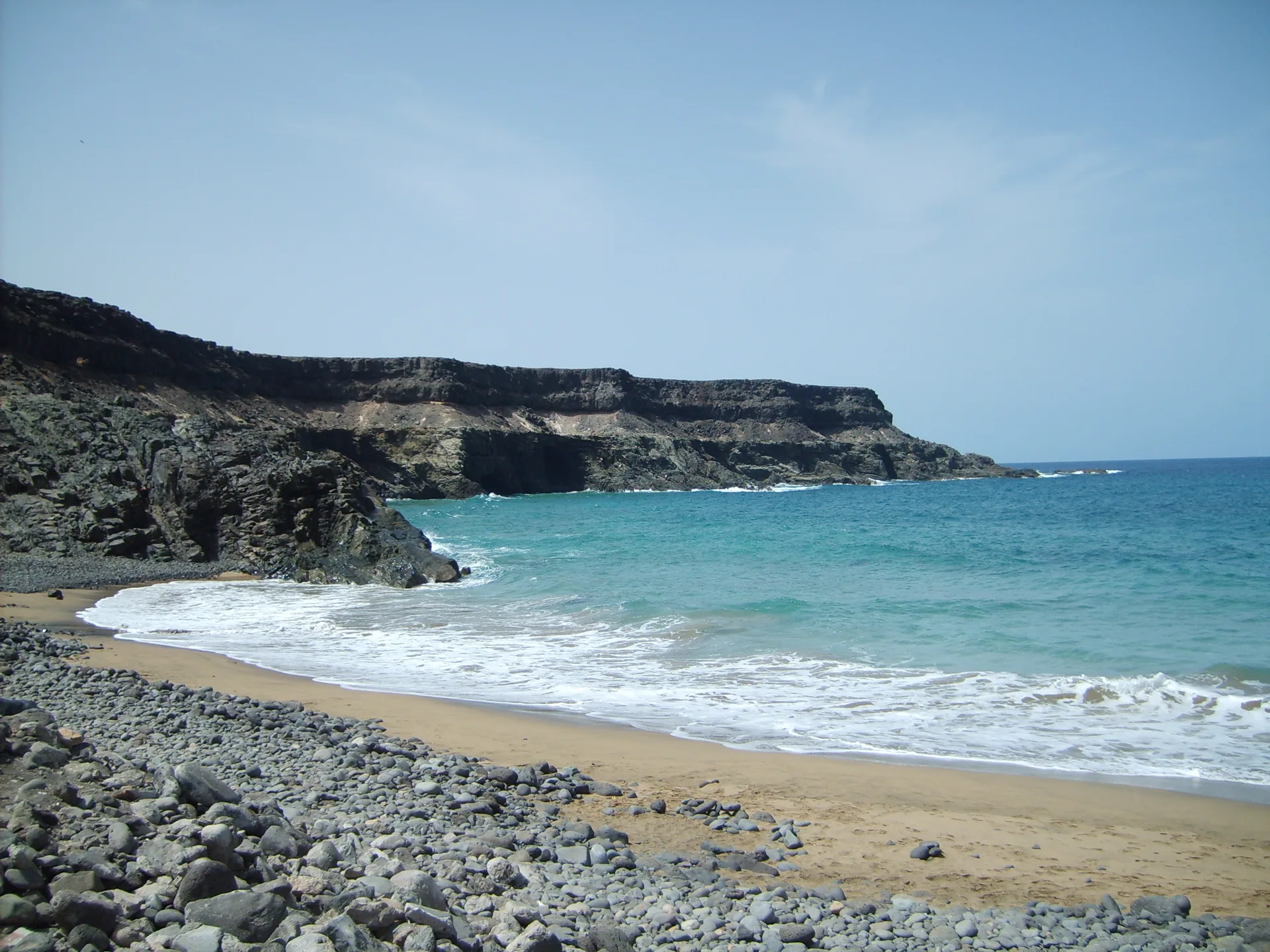 A rocky cove beach at Puertito de los Molinos with dark volcanic cliffs, a sea cave in the cliff face and turquoise water