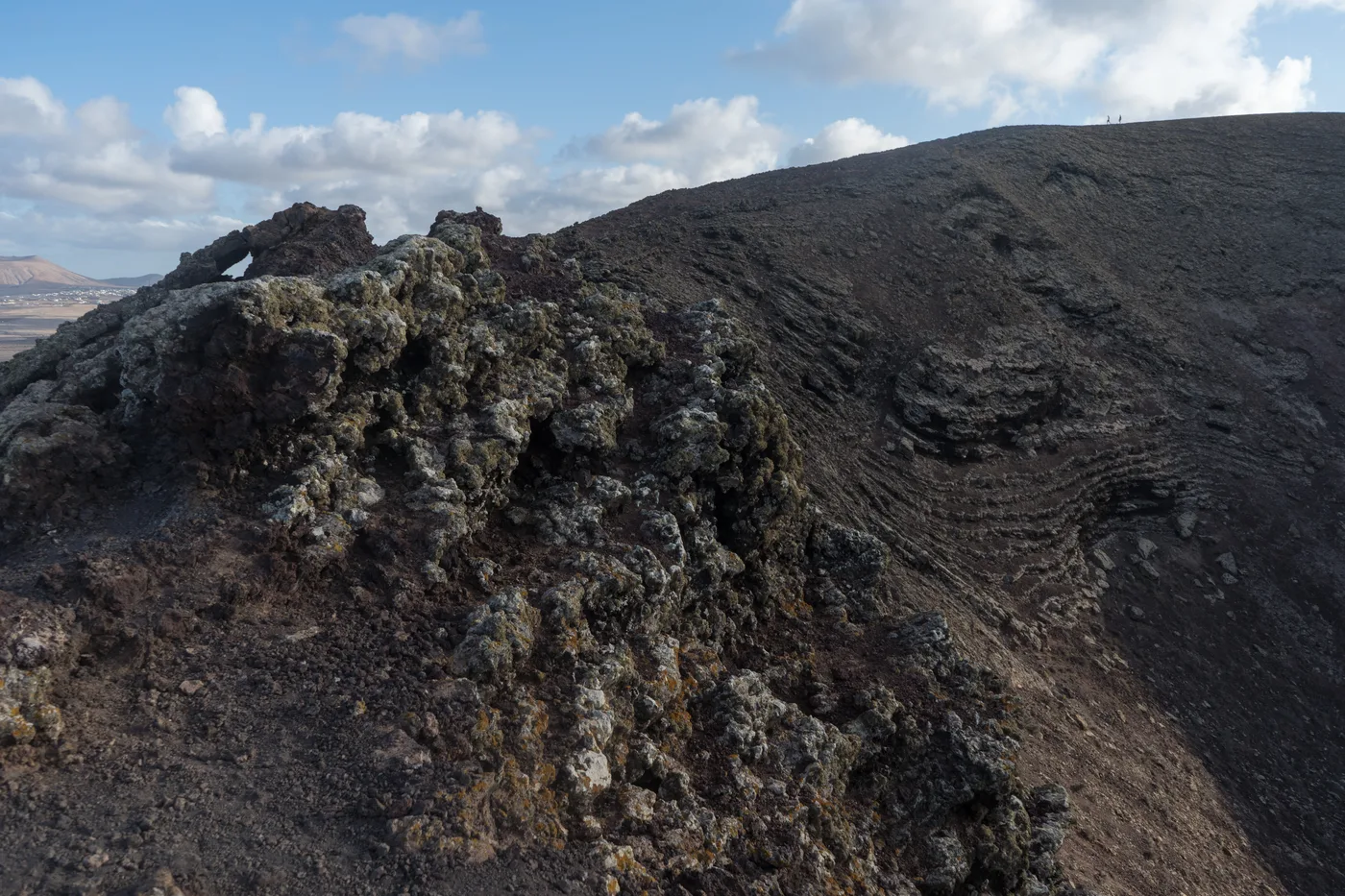 The volcanic crater rim of Calderón Hondo, its dark rock ridges leading up to the summit with a village visible on the plain below