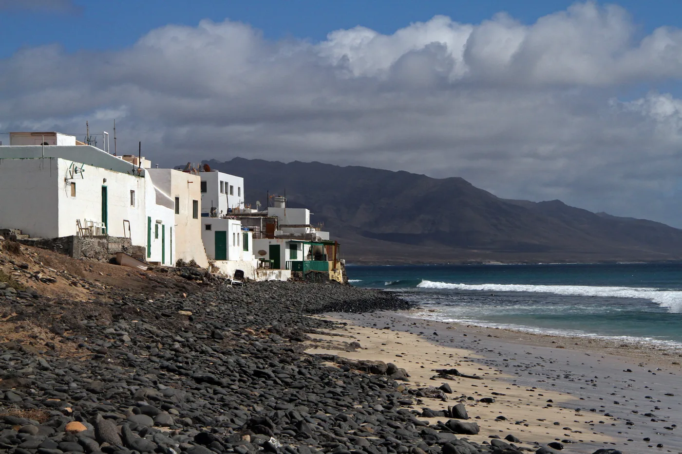 The small whitewashed fishing hamlet of El Puertito on Fuerteventura's remote west coast, its houses set above a dark volcanic-stone beach with waves breaking and bare mountains behind