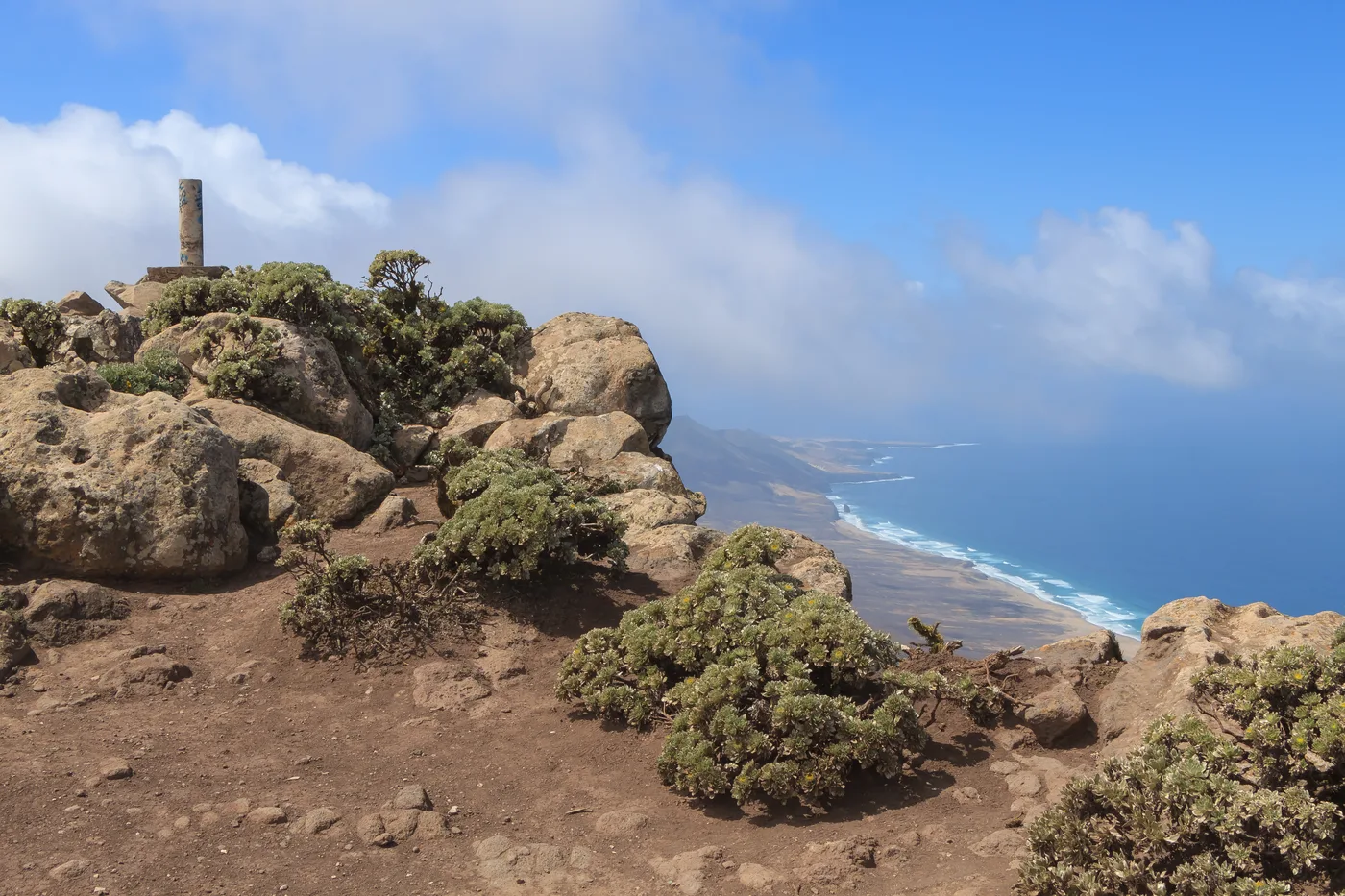 The rocky, wind-scoured summit of Pico de la Zarza, marked by an old post among boulders and low scrub, with a sweeping view down the Jandía coastline to a distant beach and the sea